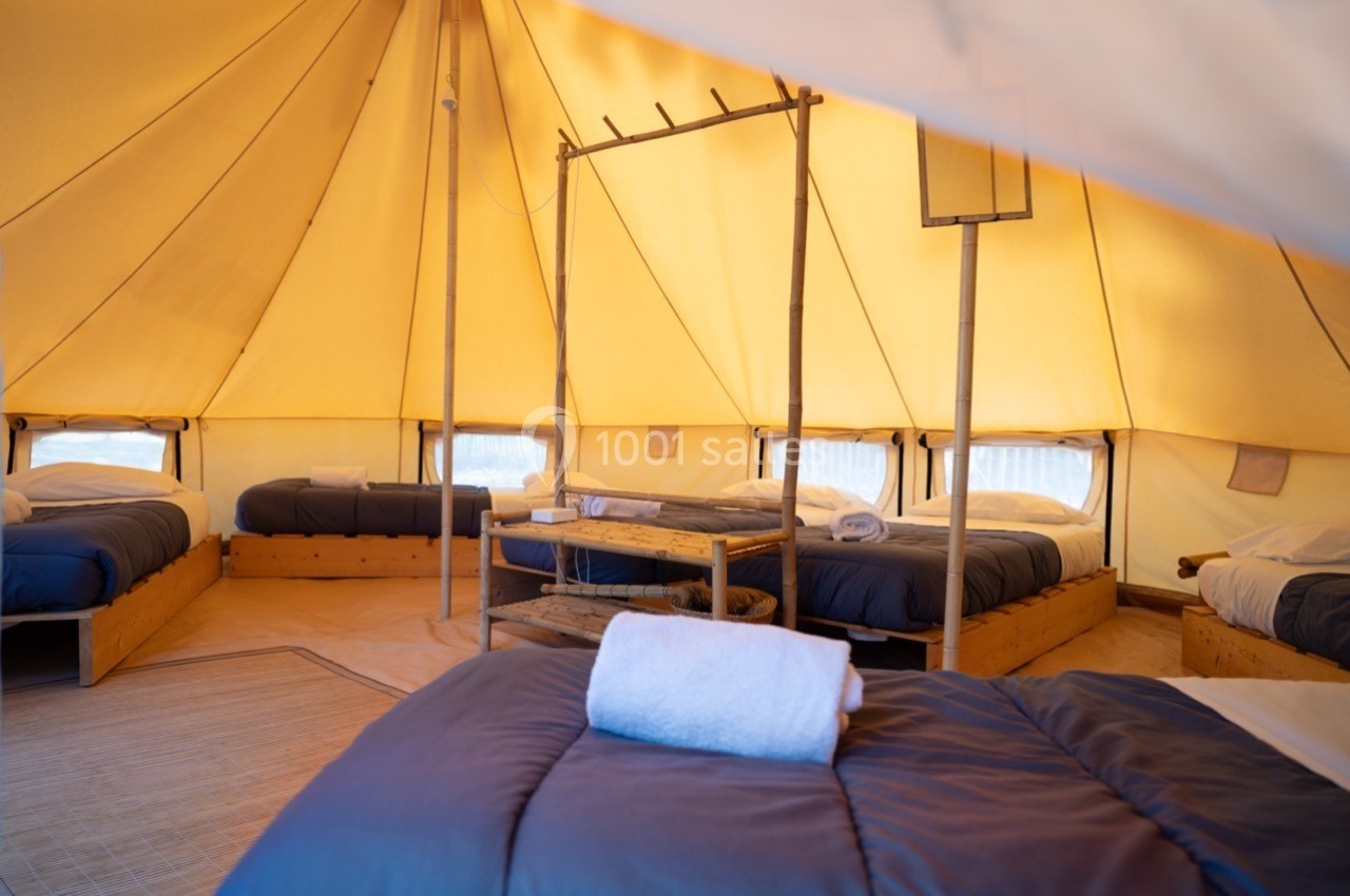 Intérieur d'une tente spacieuse avec plusieurs lits simples, draps bleus et serviettes blanches, éclairée par une lumière…