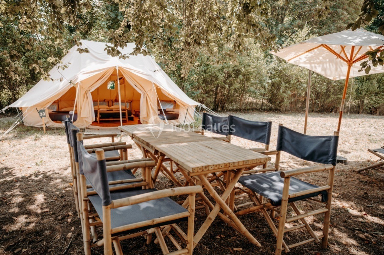 Tente aménagée avec table et chaises en bois sous des arbres, dans un cadre naturel ensoleillé.