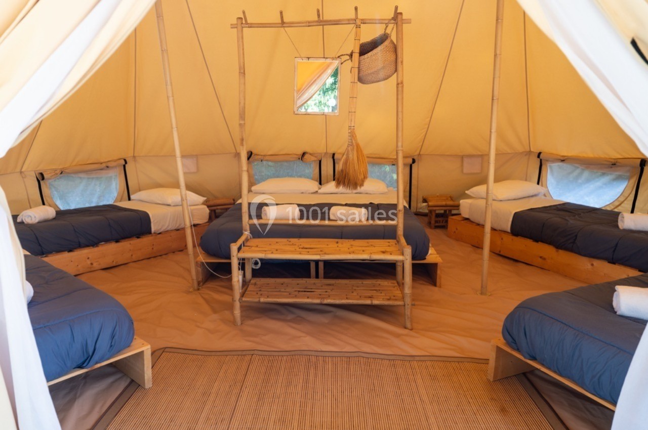 Intérieur d'une tente spacieuse avec cinq lits simples, draps bleus et mobilier en bois, éclairée par une lumière naturelle.