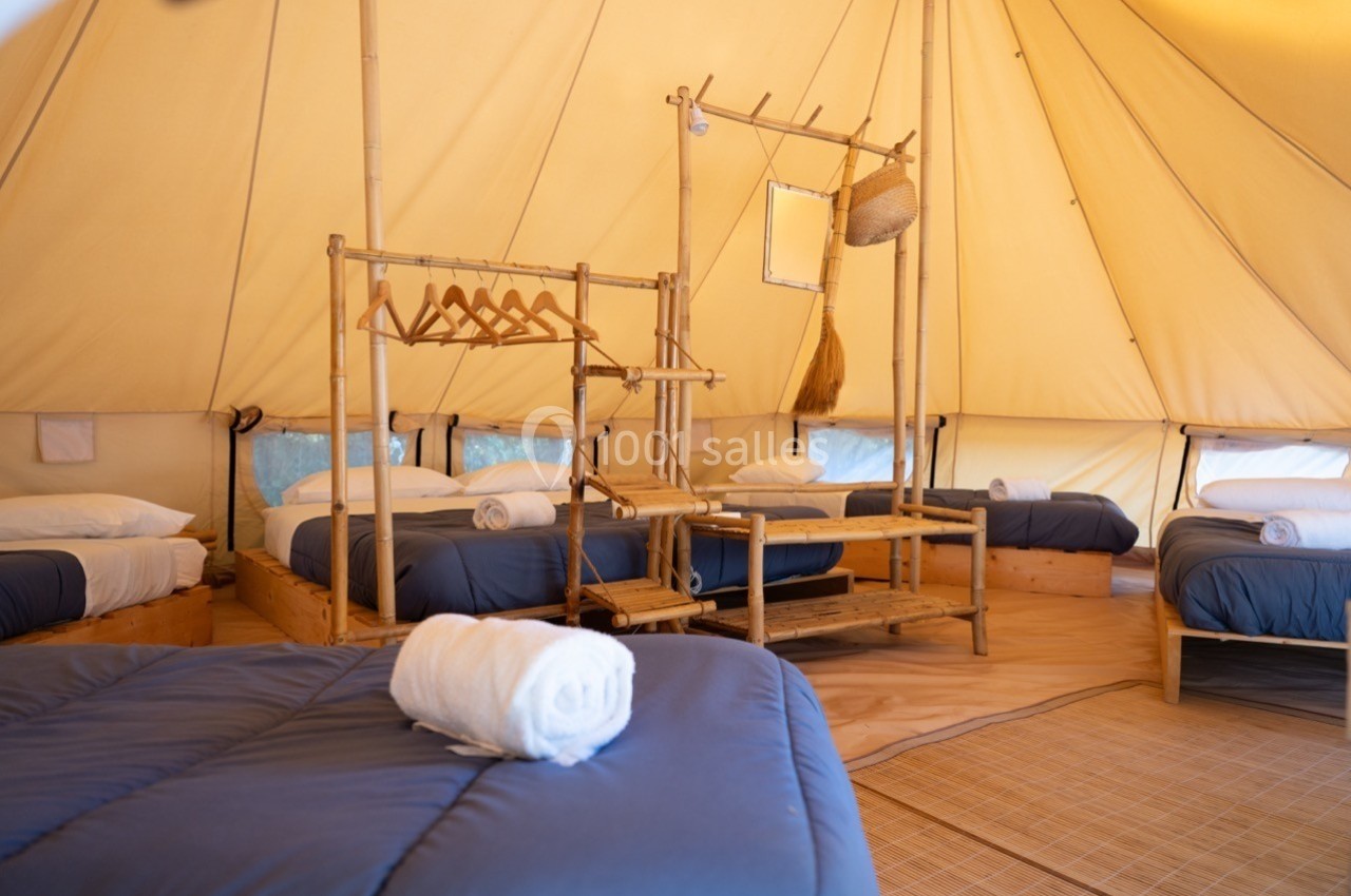 Intérieur d'une tente spacieuse avec lits simples, draps bleus, serviettes roulées et mobilier en bambou.