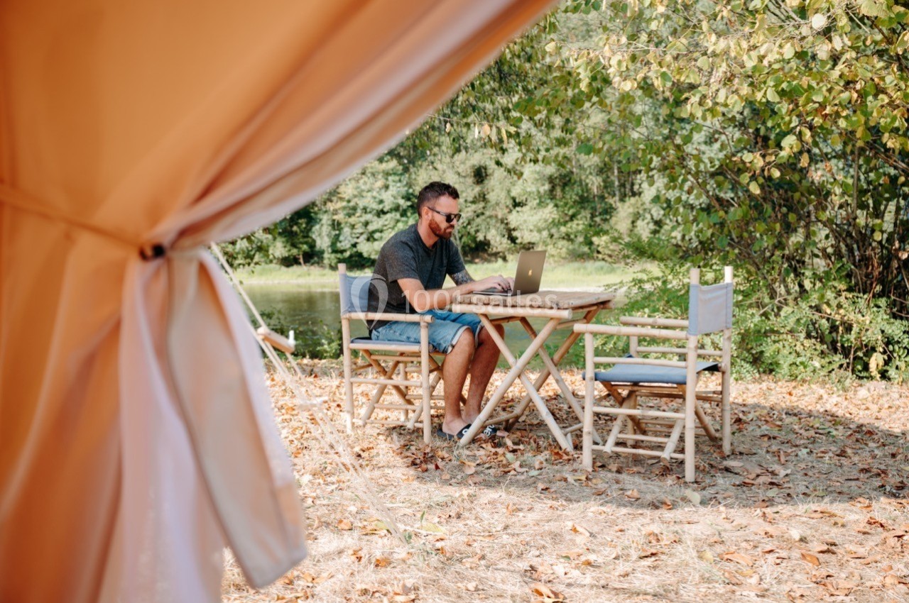 Un homme travaille sur un ordinateur portable à une table en plein air, entouré de nature et de feuilles au sol.