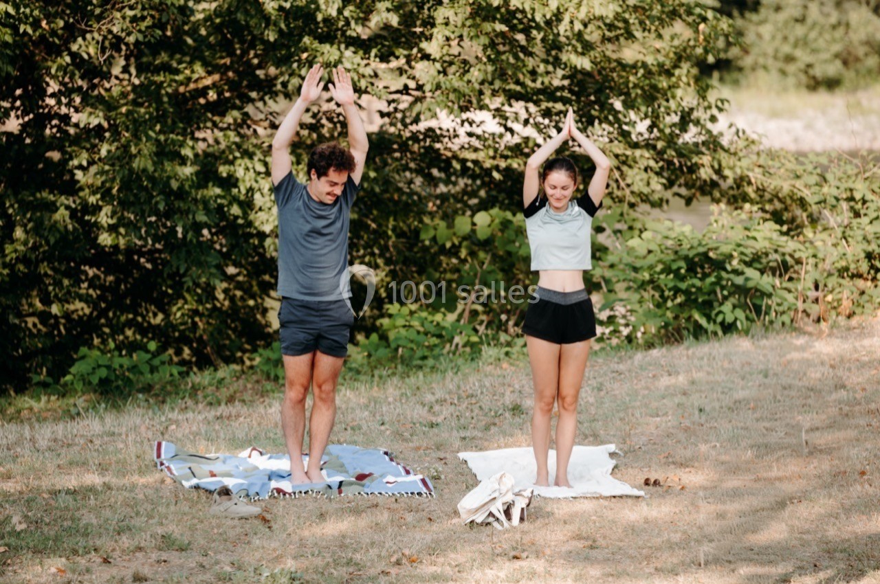 Deux personnes pratiquent le yoga en plein air sur des tapis, entourées de végétation.