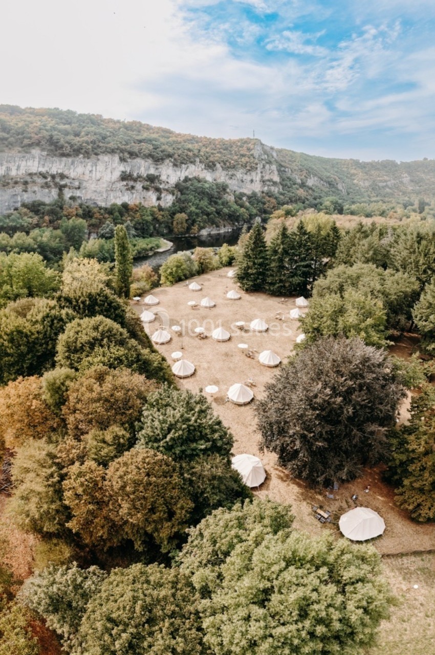 Vue aérienne d'un terrain boisé avec des tentes blanches dispersées, entouré de collines et d'arbres.