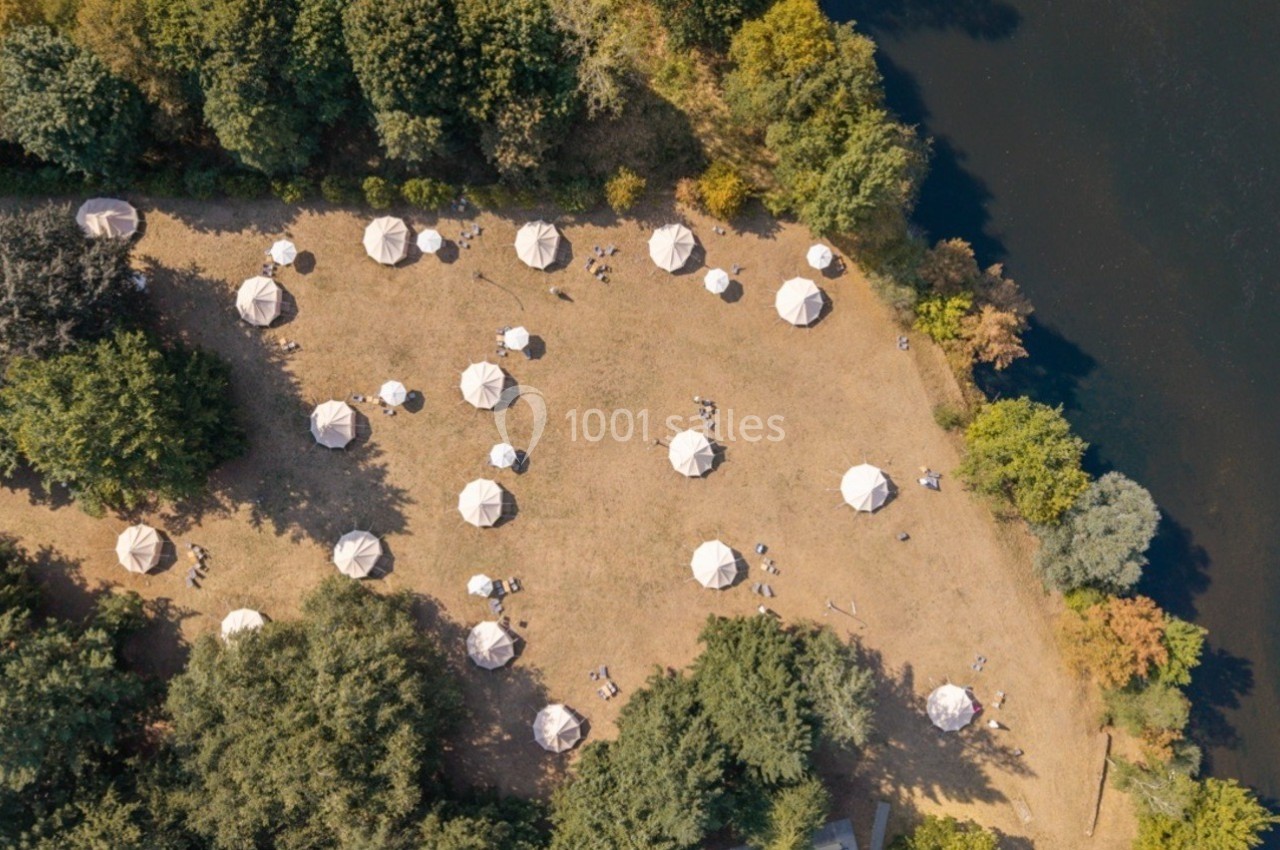 Vue aérienne d'un terrain herbeux avec des tentes blanches disposées en cercle près d'une rivière bordée d'arbres.