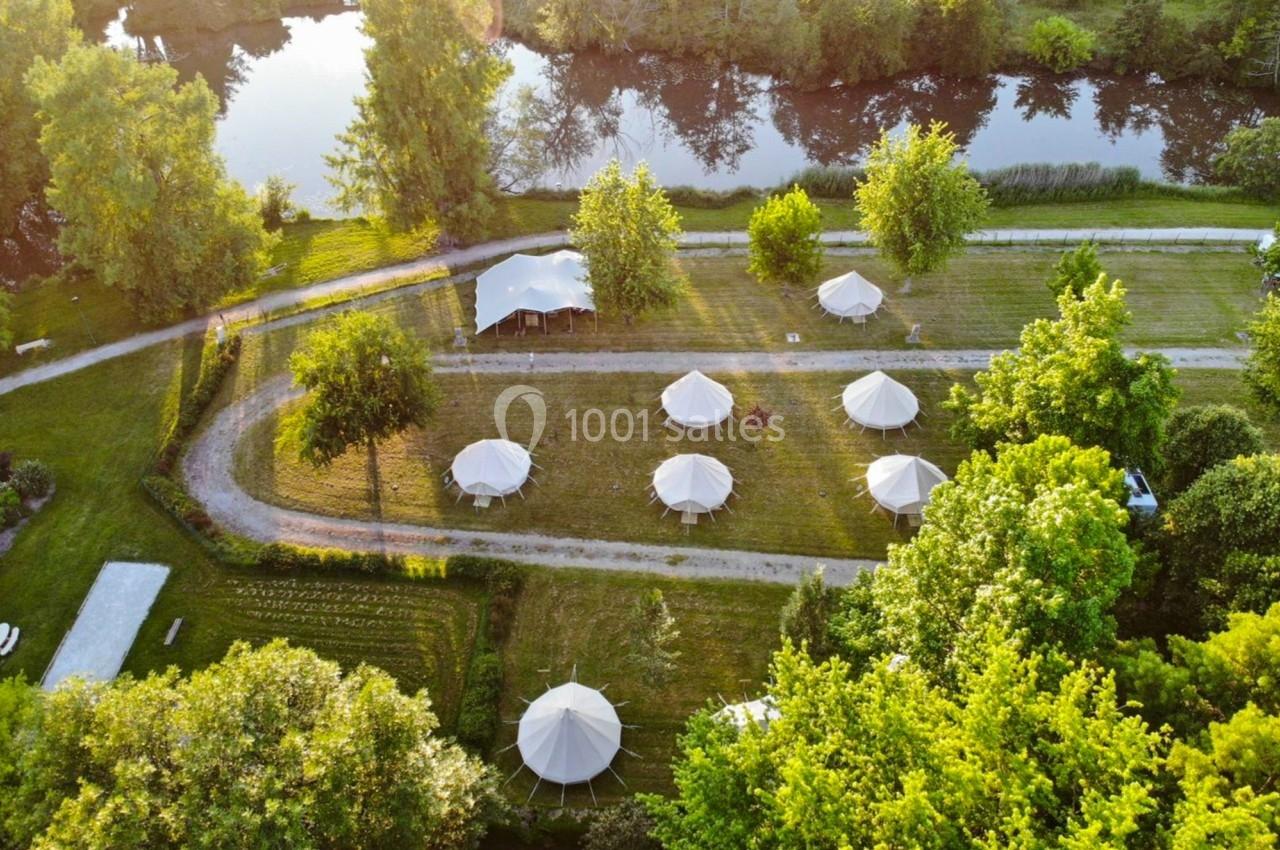 Vue aérienne d'un site de glamping avec des tentes blanches disposées près d'un chemin entouré de verdure et d'un plan d'eau.