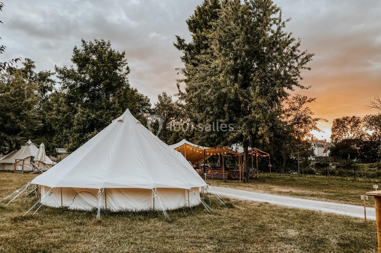 Tente blanche installée sur une pelouse, entourée d'arbres et de structures en toile, au coucher du soleil.