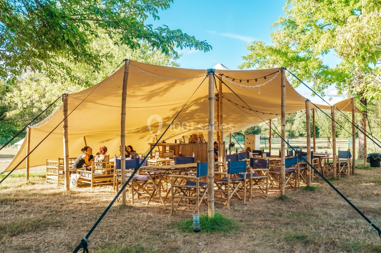 Tente en toile beige montée en extérieur, abritant des tables et des chaises en bois, entourée d'arbres.