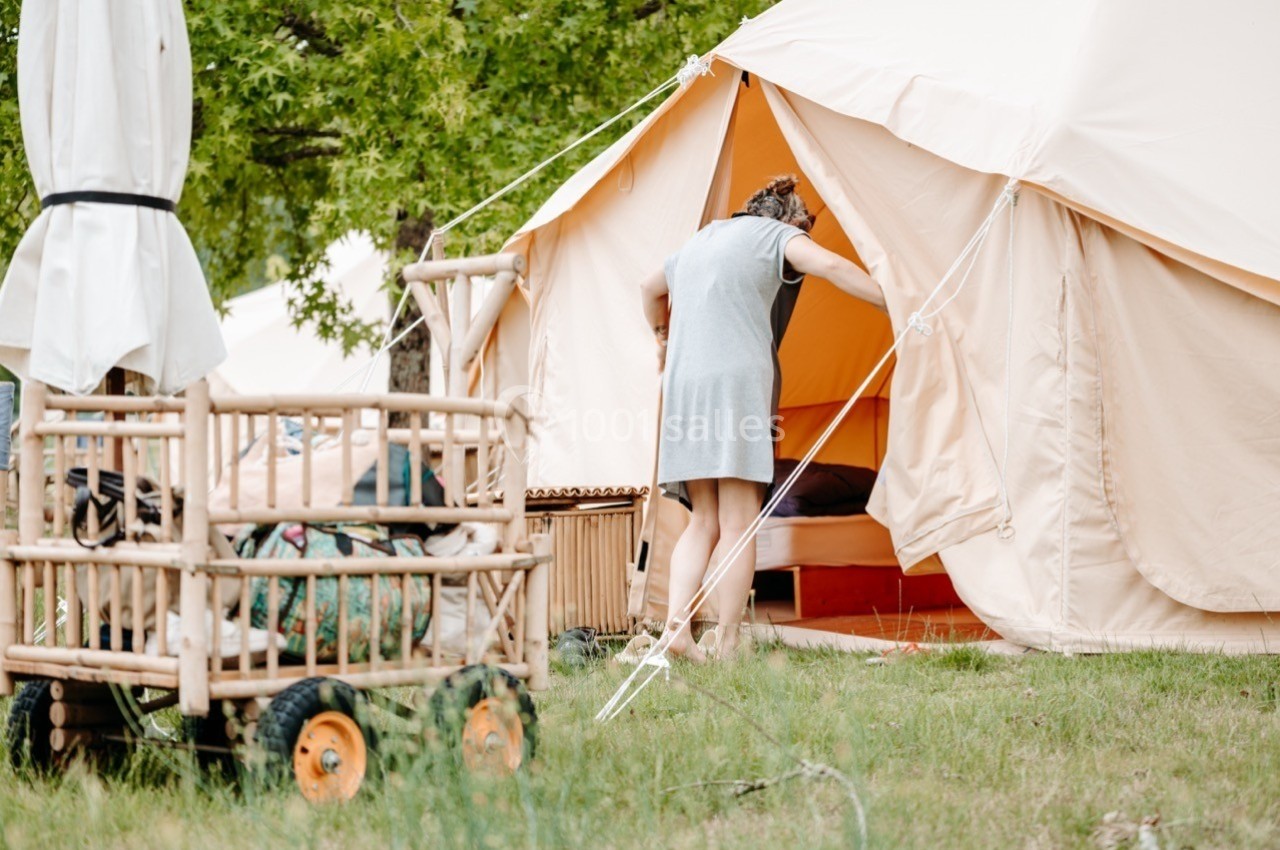 Une femme entre dans une tente beige installée sur une pelouse, avec un chariot en bois chargé d'affaires à proximité.