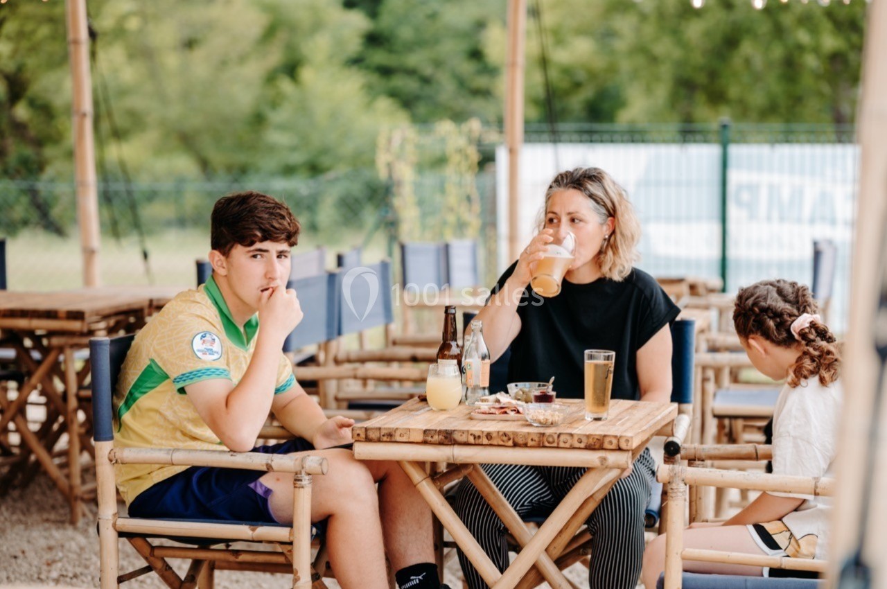 Deux personnes et un enfant assis à une table en bois, mangeant et buvant en extérieur, avec un fond verdoyant.
