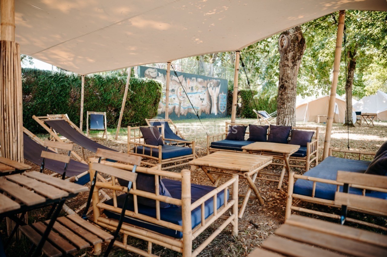 Mobilier en bambou avec coussins bleus disposé sous une toile d'ombrage dans un espace extérieur arboré.