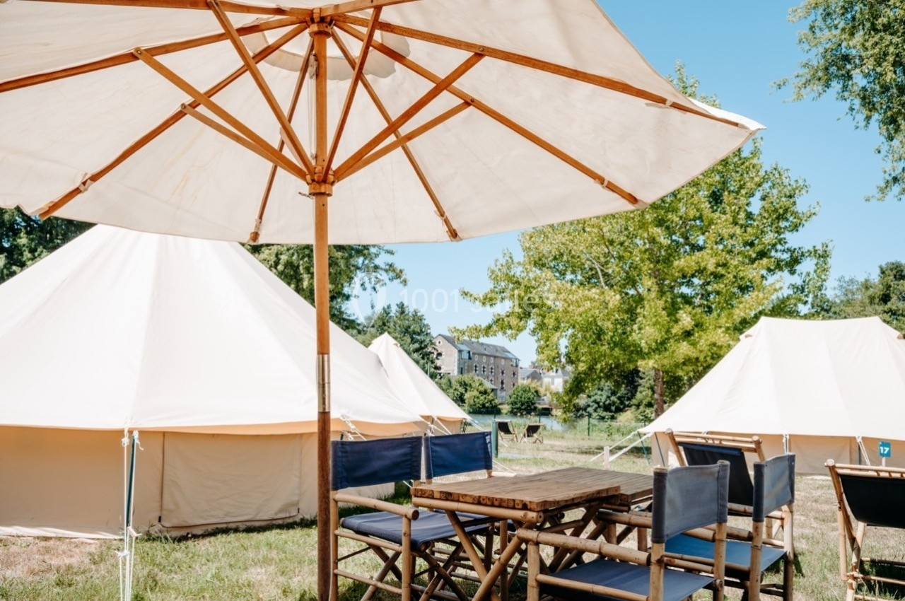 Table et chaises en bois sous un parasol, entourées de tentes blanches dans un espace extérieur verdoyant.