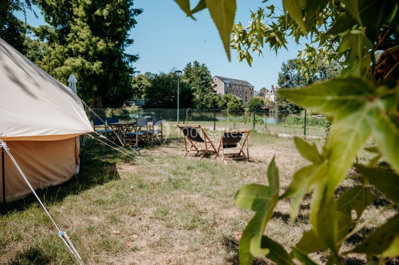 Tente beige, chaises longues et table en extérieur sur une pelouse, avec arbres et bâtiment en arrière-plan.