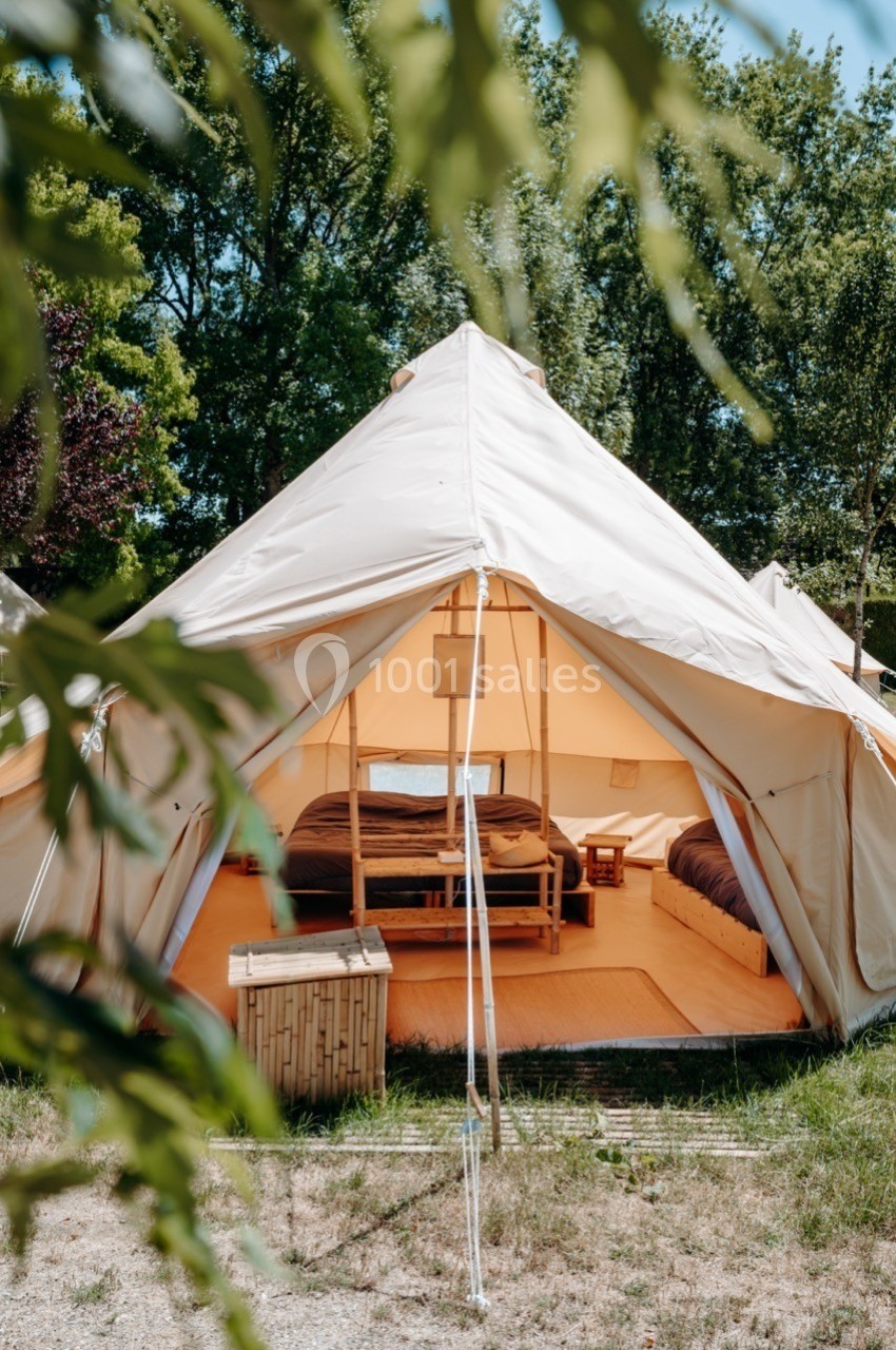 Tente glamping beige avec intérieur aménagé, comprenant un lit double et du mobilier en bois, entourée de verdure.