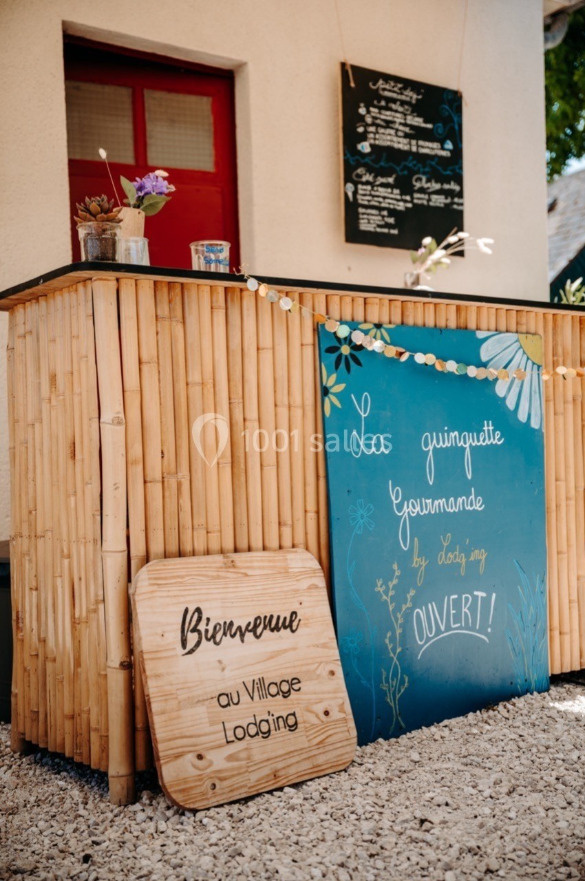 Comptoir en bambou avec panneaux indiquant ’La guinguette gourmande’ et ’Bienvenue’, devant une façade claire.