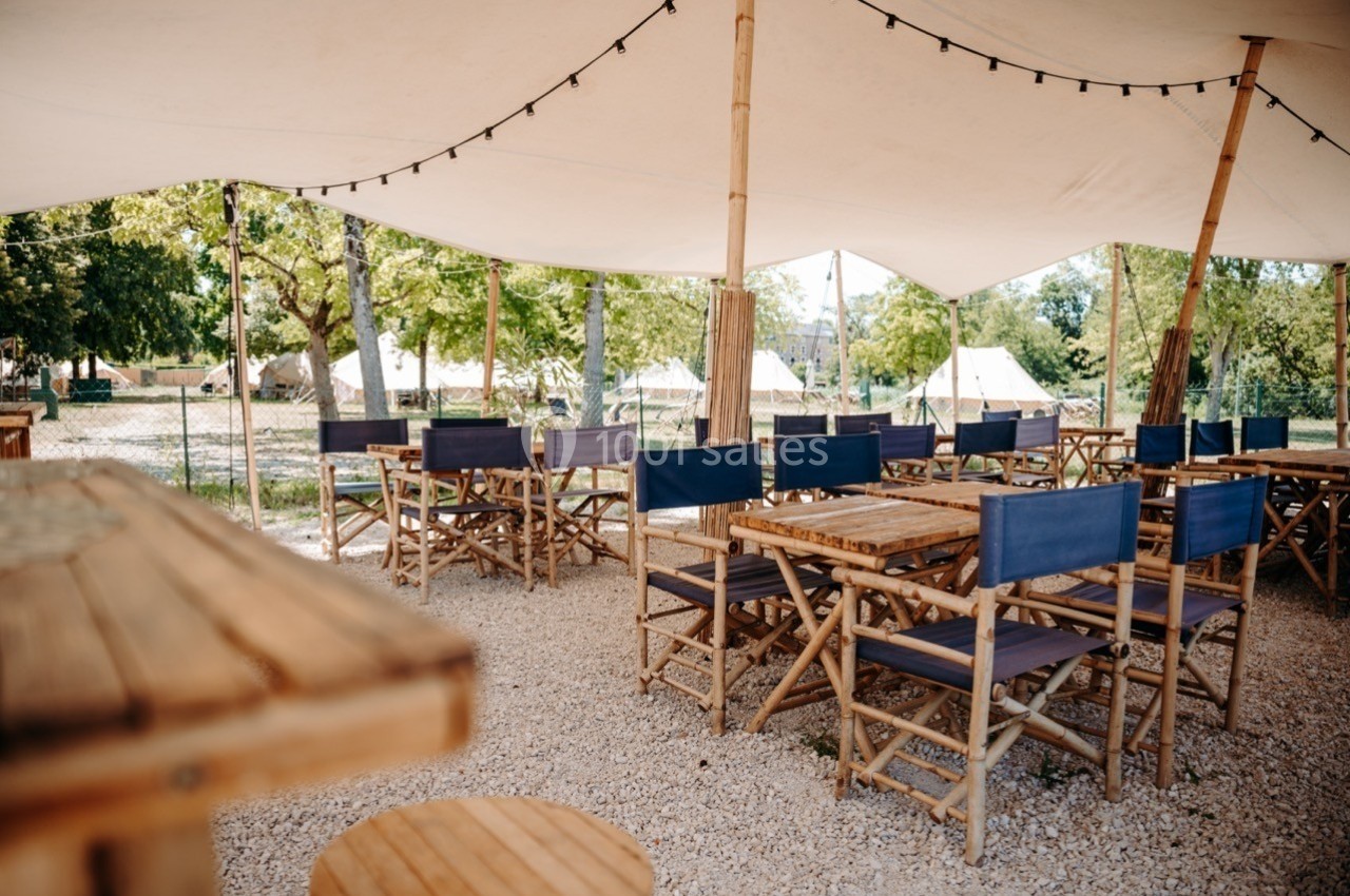 Terrasse extérieure avec tables et chaises en bois sous une toile blanche, entourée d'arbres et de lumière naturelle.