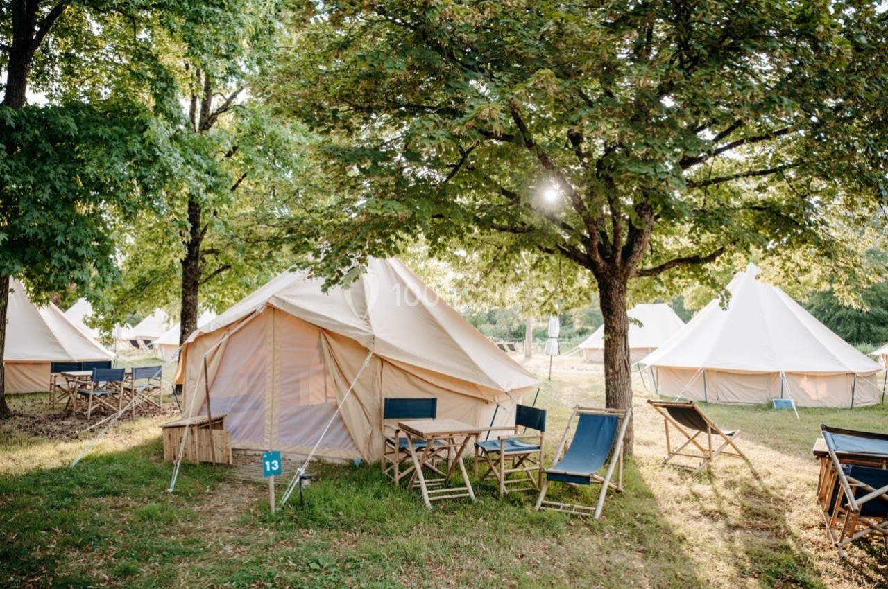 Tentes en toile beige installées sous des arbres, entourées de chaises et de tables en bois sur une pelouse.