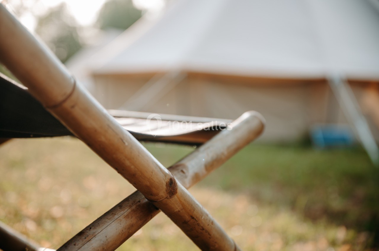 Chaise pliante en bambou au premier plan, avec une tente floue en arrière-plan dans un cadre extérieur verdoyant.