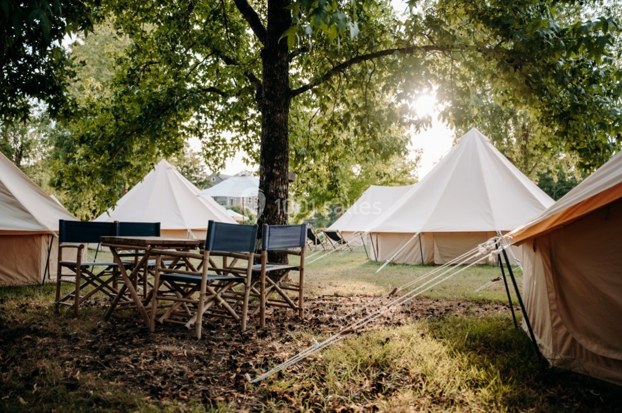 Tentes en toile beige installées sous des arbres, avec une table et des chaises en bois au premier plan.