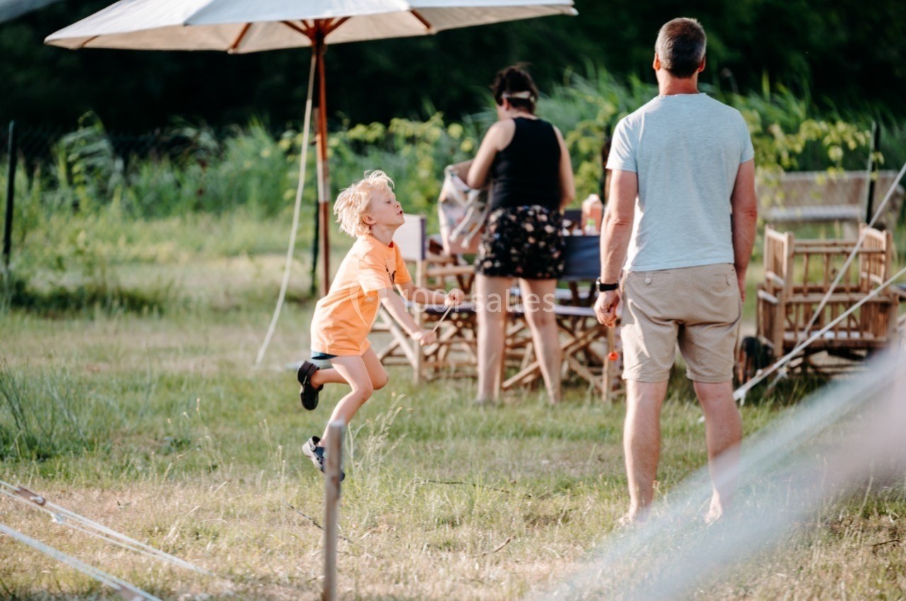 Un enfant saute près d'un homme debout, avec une table et des chaises en bois en arrière-plan dans un jardin.