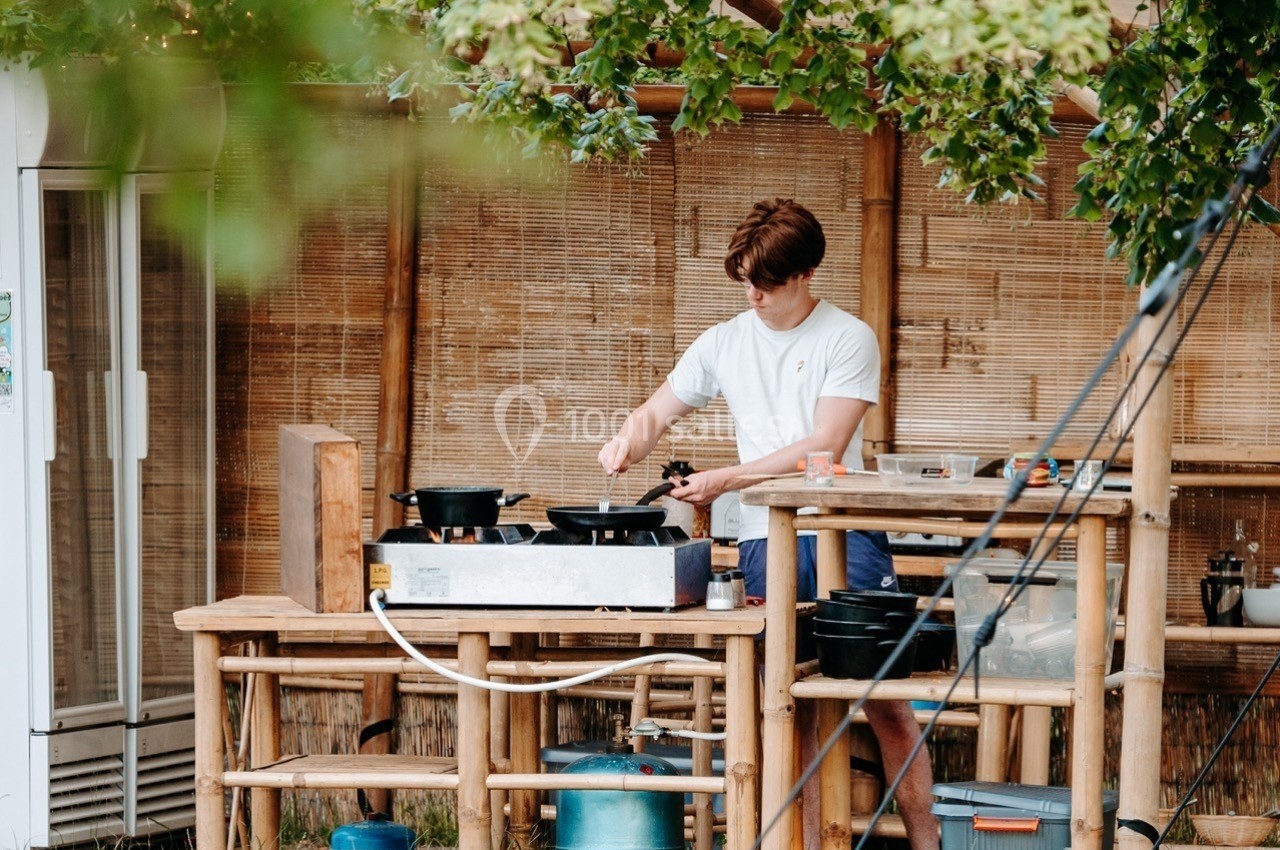 Un homme cuisine sur une plaque chauffante dans une cuisine extérieure en bois, entourée de végétation.