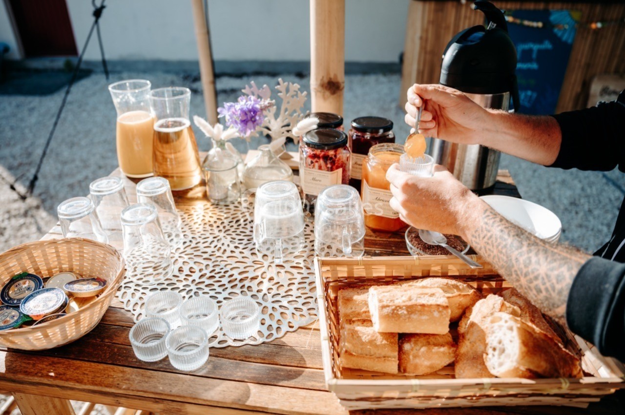 Table en bois avec verres, jus, confitures, pain et une personne servant du miel dans un verre.
