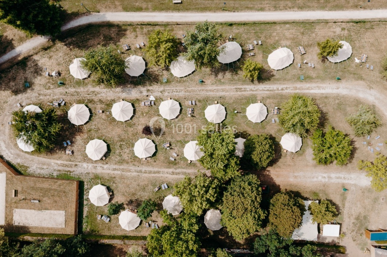 Vue aérienne d'un espace extérieur avec des parasols blancs alignés, entourés de verdure et de chemins en terre.