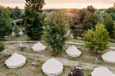 Miniature Location salle Cellettes (Loir-et-Cher) - Lodg’ing Nature Camp Châteaux de la Loire #24 Tentes blanches installées dans un espace naturel près d'une rivière, entourées d'arbres et d'un pont au coucher du soleil.