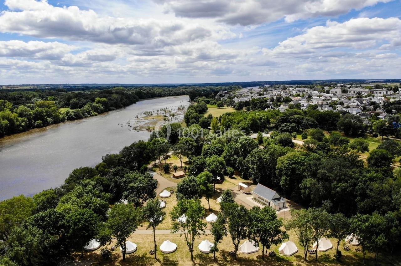 Vue aérienne d'un camping avec des tentes blanches entourées d'arbres, près d'une rivière et d'un village.