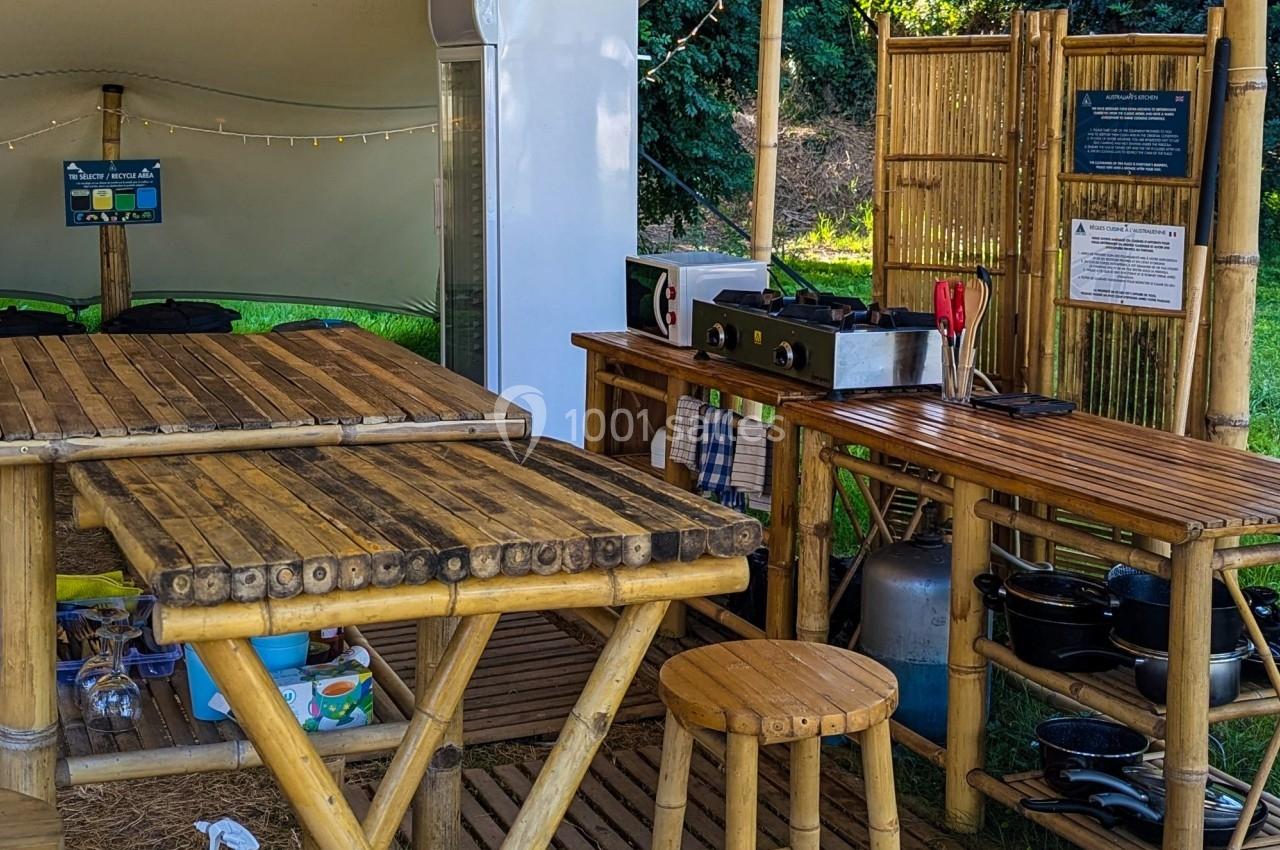 Cuisine extérieure en bambou avec plan de travail, tabouret, réfrigérateur, ustensiles et vaisselle dans un cadre naturel.