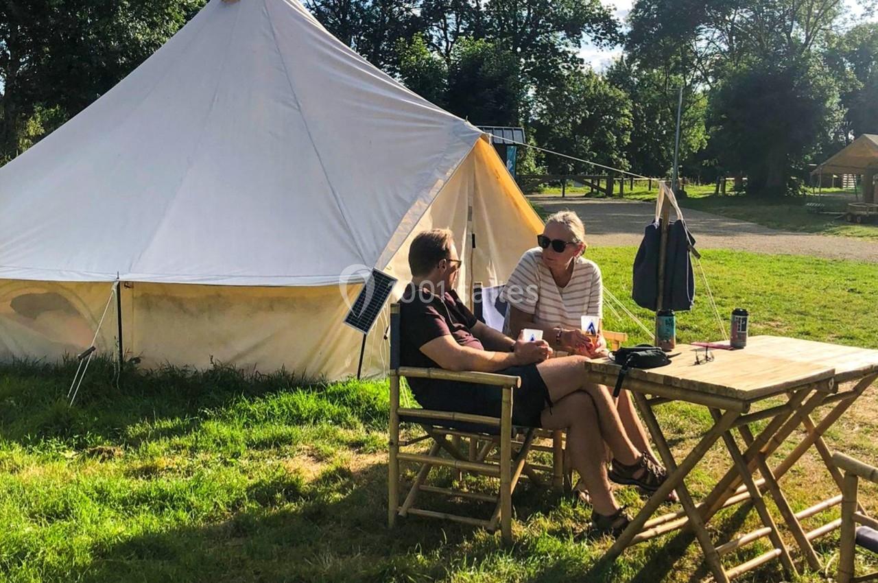 Deux personnes assises à une table en bois devant une tente blanche dans un camping ensoleillé.