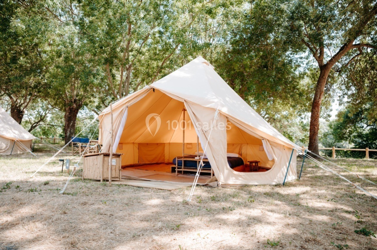 Tente en toile beige aménagée avec un lit, située dans une clairière entourée d'arbres.