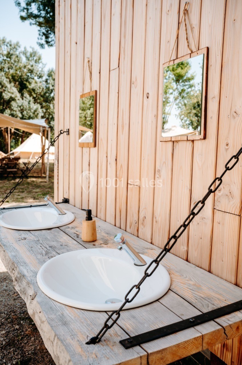 Deux lavabos extérieurs avec miroirs suspendus sur un mur en bois, entourés de végétation et de tentes en arrière-plan.