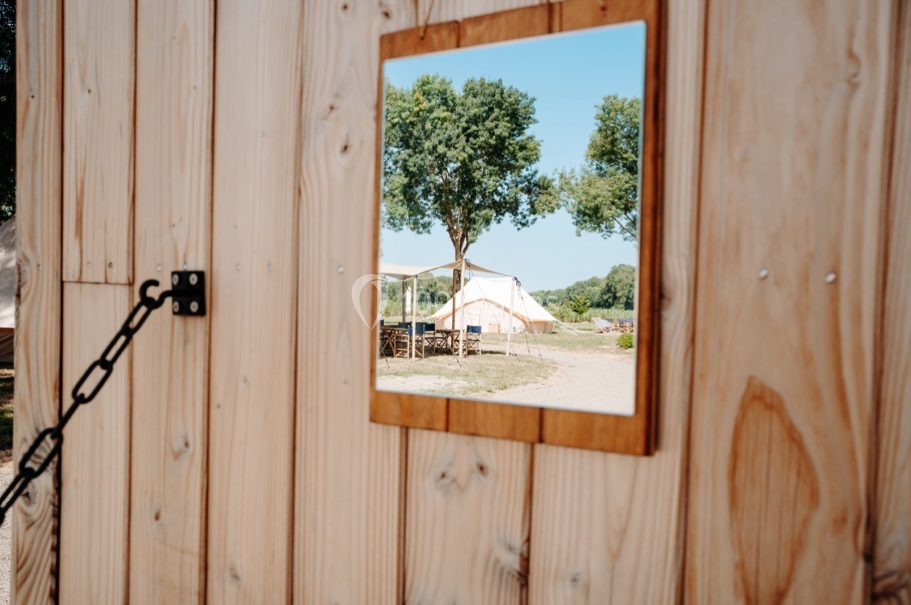 Reflet d'un paysage avec arbres et tente blanche dans un miroir encadré en bois fixé sur une paroi en bois clair.