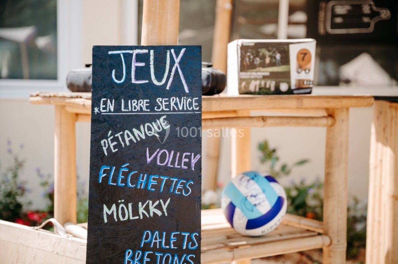 Panneau en ardoise listant des jeux en libre-service, avec un ballon de volley posé sur une table en bois.