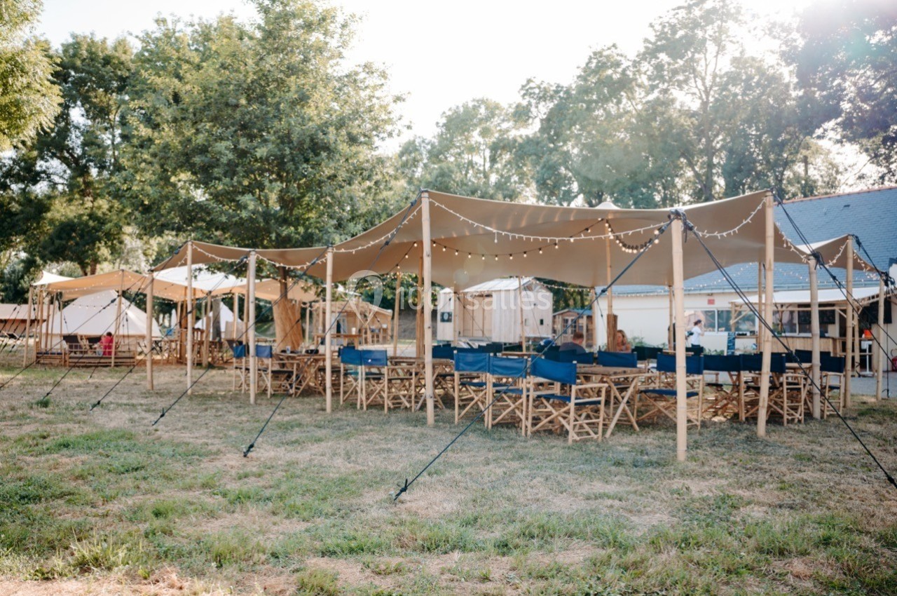 Tente beige avec des guirlandes lumineuses, chaises pliantes et tables en bois sur une pelouse entourée d'arbres.