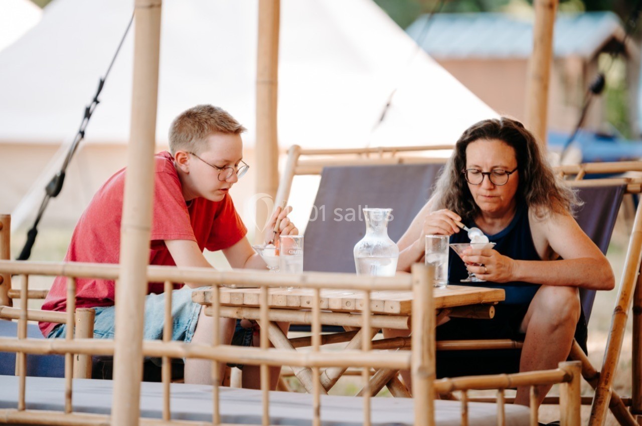 Deux personnes assises à une table en bambou, mangeant et buvant, dans un cadre extérieur avec des tentes en arrière-plan.