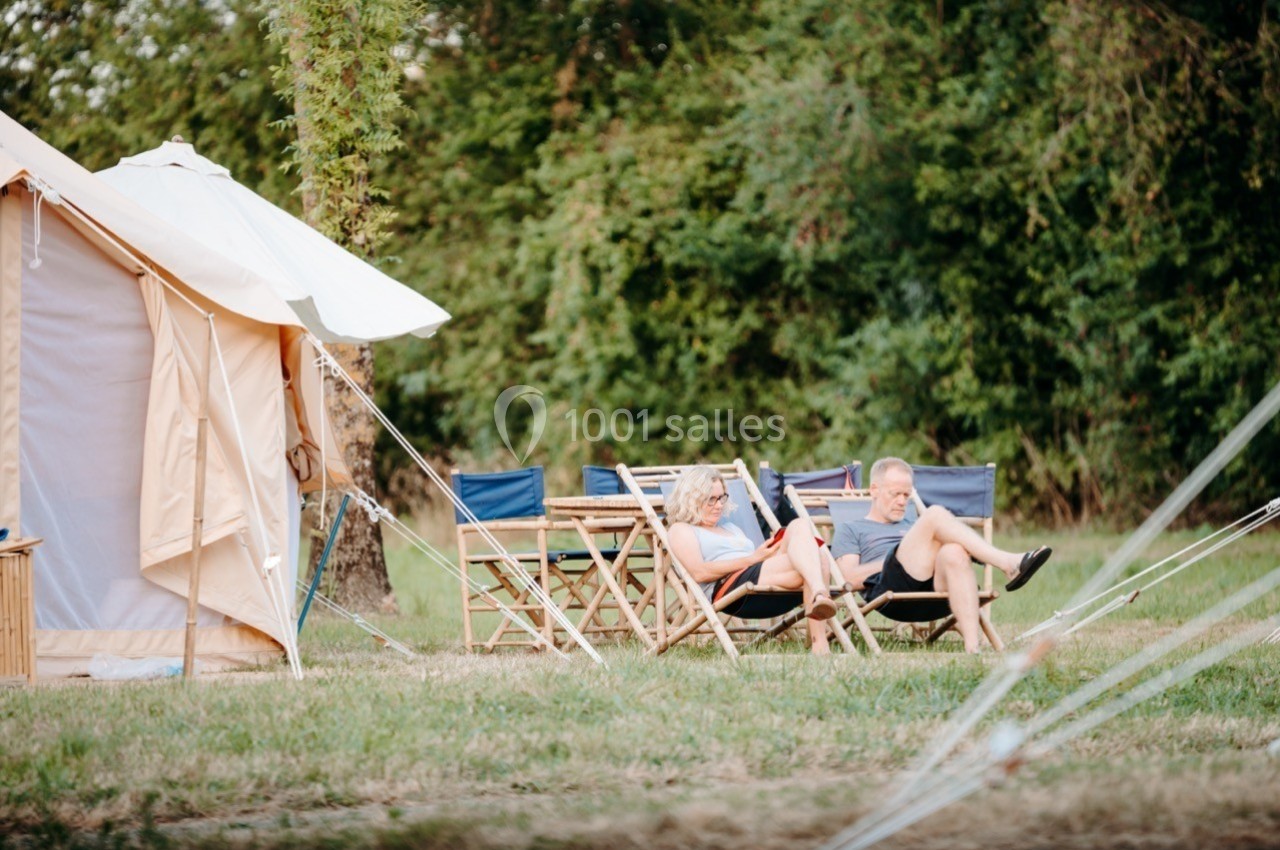 Deux personnes assises sur des chaises pliantes devant une tente, dans un cadre naturel avec des arbres en arrière-plan.