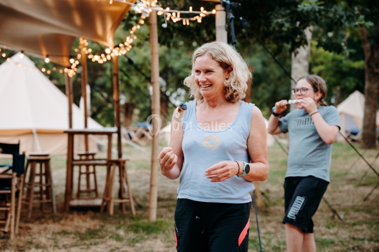 Une femme souriante marche dans un espace extérieur avec des tentes et des guirlandes lumineuses en arrière-plan.