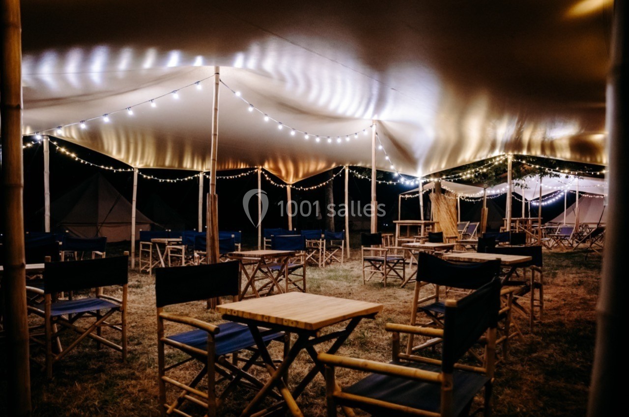 Chaises et tables en bois disposées sous une tente éclairée par des guirlandes lumineuses dans un cadre extérieur nocturne.