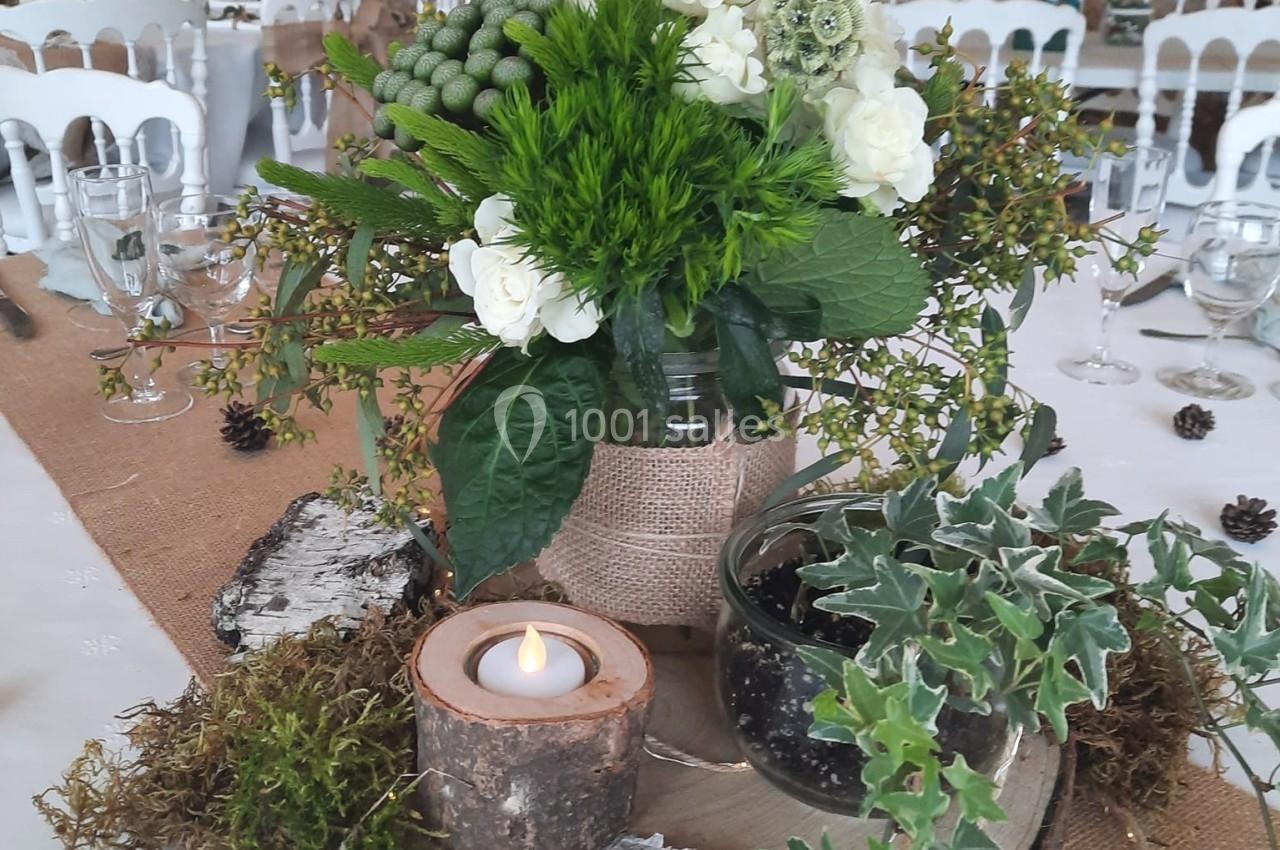 Centre de table rustique avec fleurs blanches, feuillage, bougie allumée et numéro ’Quatre’ sur fond de salle en pierre.