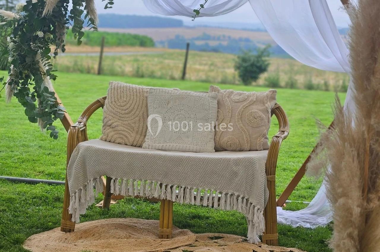 Banc en rotin décoré de coussins et d'un plaid, sous une arche ornée de fleurs et de voilages, sur une pelouse.