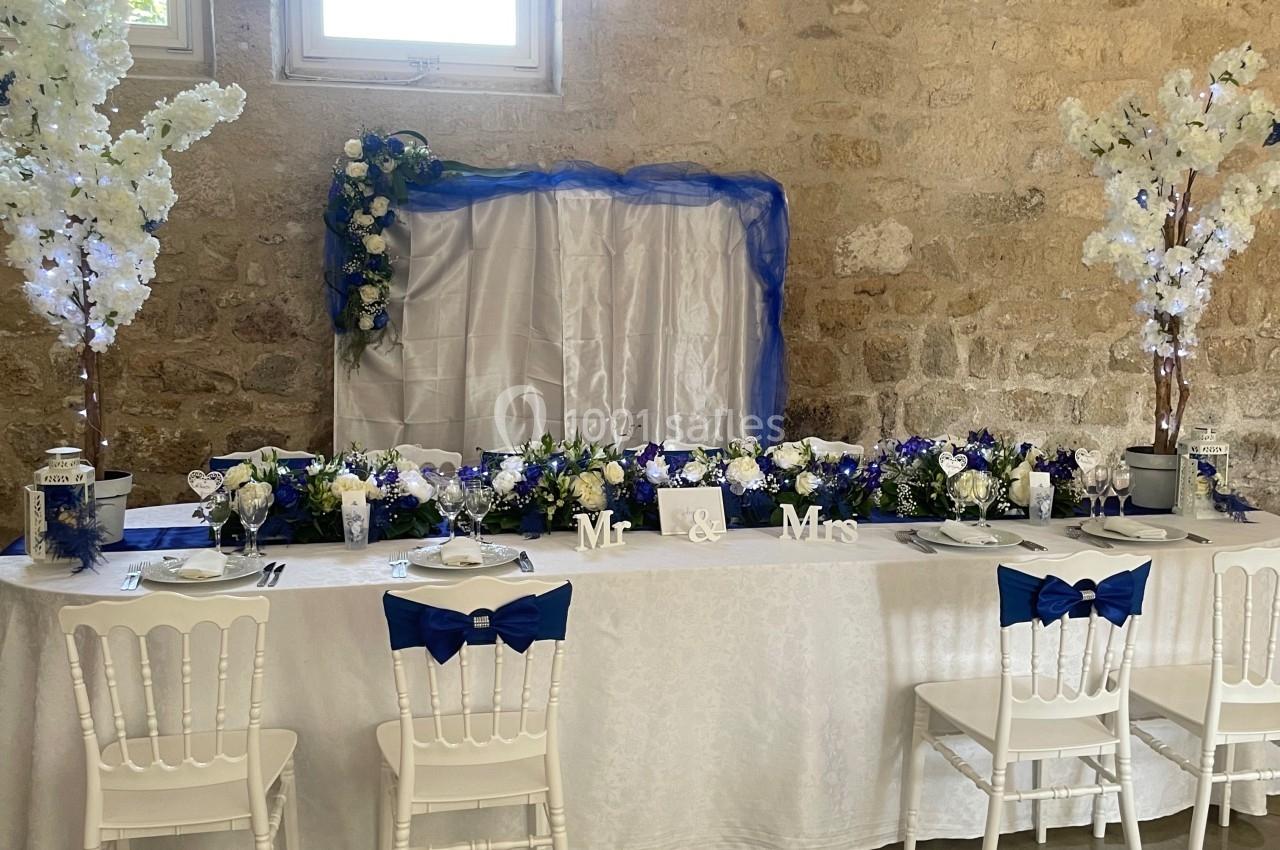 Table de mariage décorée de fleurs blanches et bleues, avec chaises blanches ornées de nœuds bleus, devant un mur en pierre.