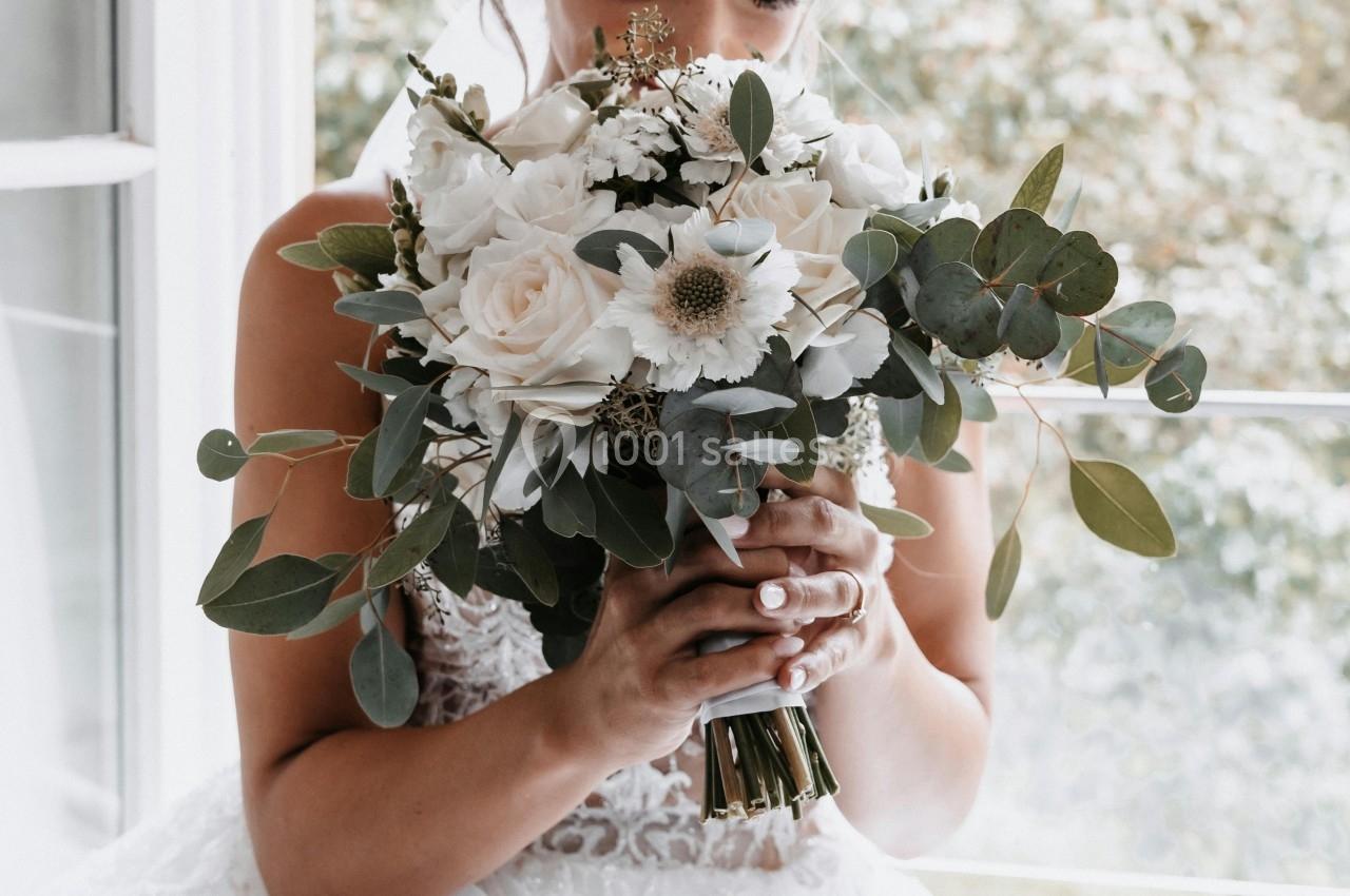Une mariée en robe blanche tient un bouquet de fleurs blanches et vertes devant une fenêtre lumineuse.