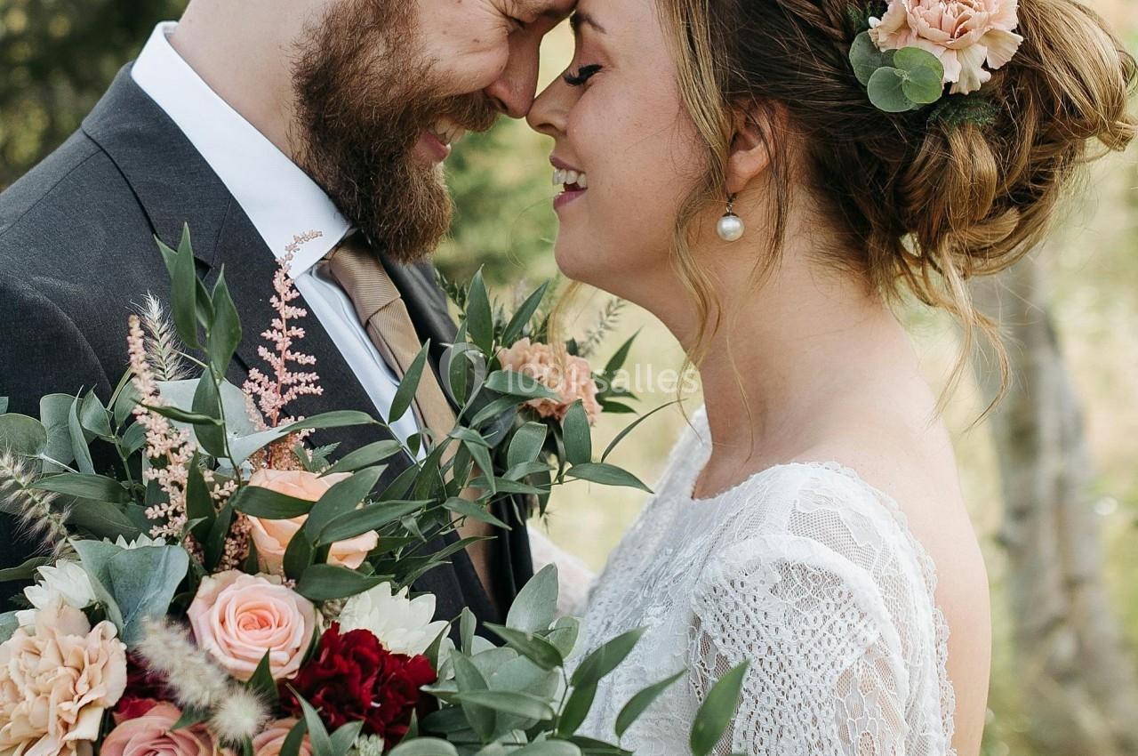 Un couple souriant en tenue de mariage se tient tête contre tête, entouré de verdure, avec un bouquet fleuri.