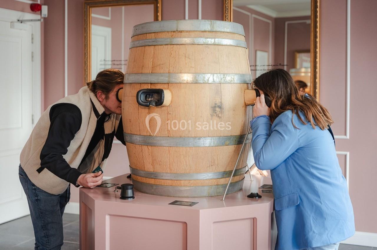Deux personnes observent à travers des oculaires fixés sur un tonneau en bois dans une pièce aux murs roses.