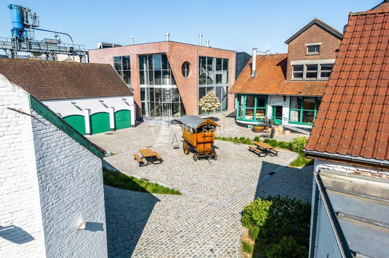 Cour pavée entourée de bâtiments en briques et en crépi, avec une roulotte en bois et des bancs au centre.