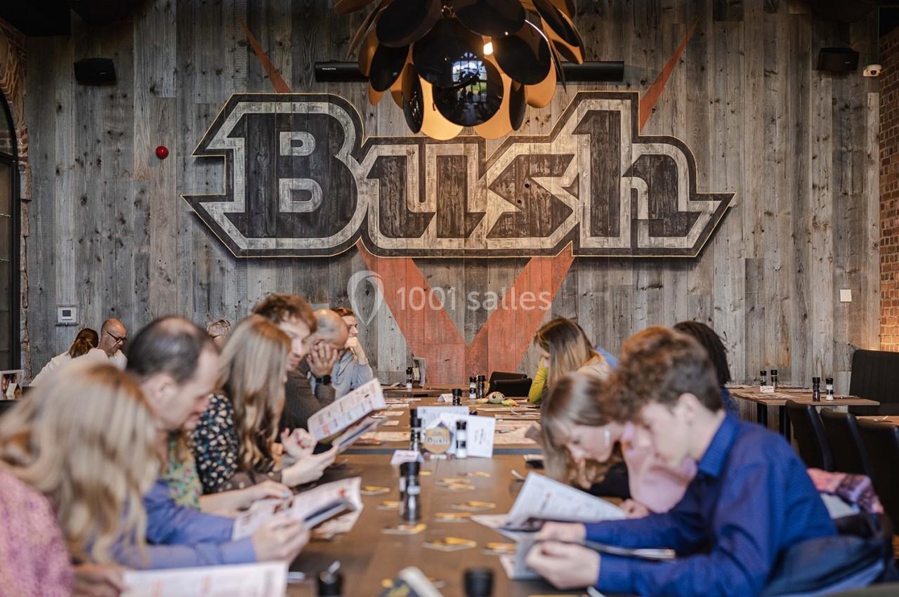 Des personnes assises autour d'une grande table dans un restaurant, lisant des menus devant un mur décoré du logo ’Bush’.