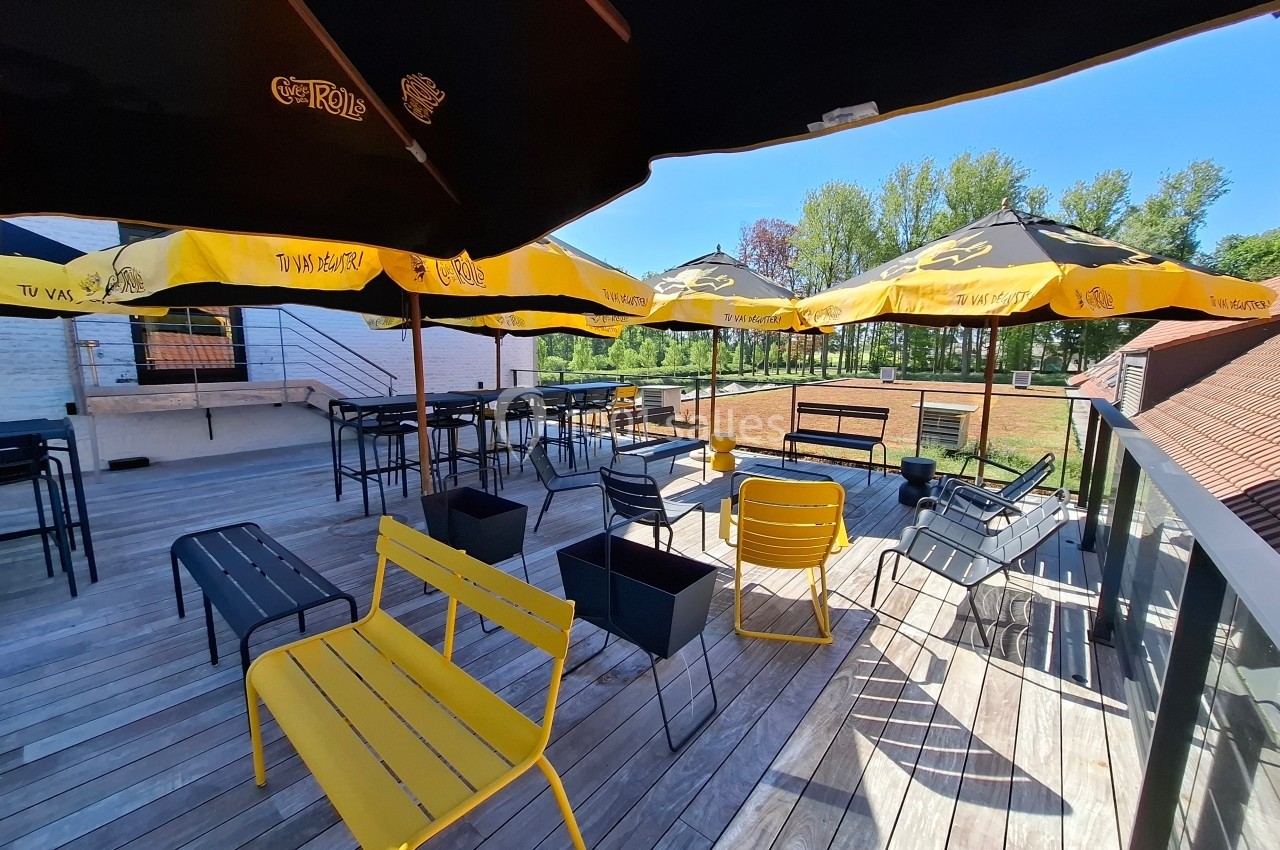 Terrasse en bois avec tables, bancs jaunes et parasols jaunes et noirs, entourée de verdure par une journée ensoleillée.