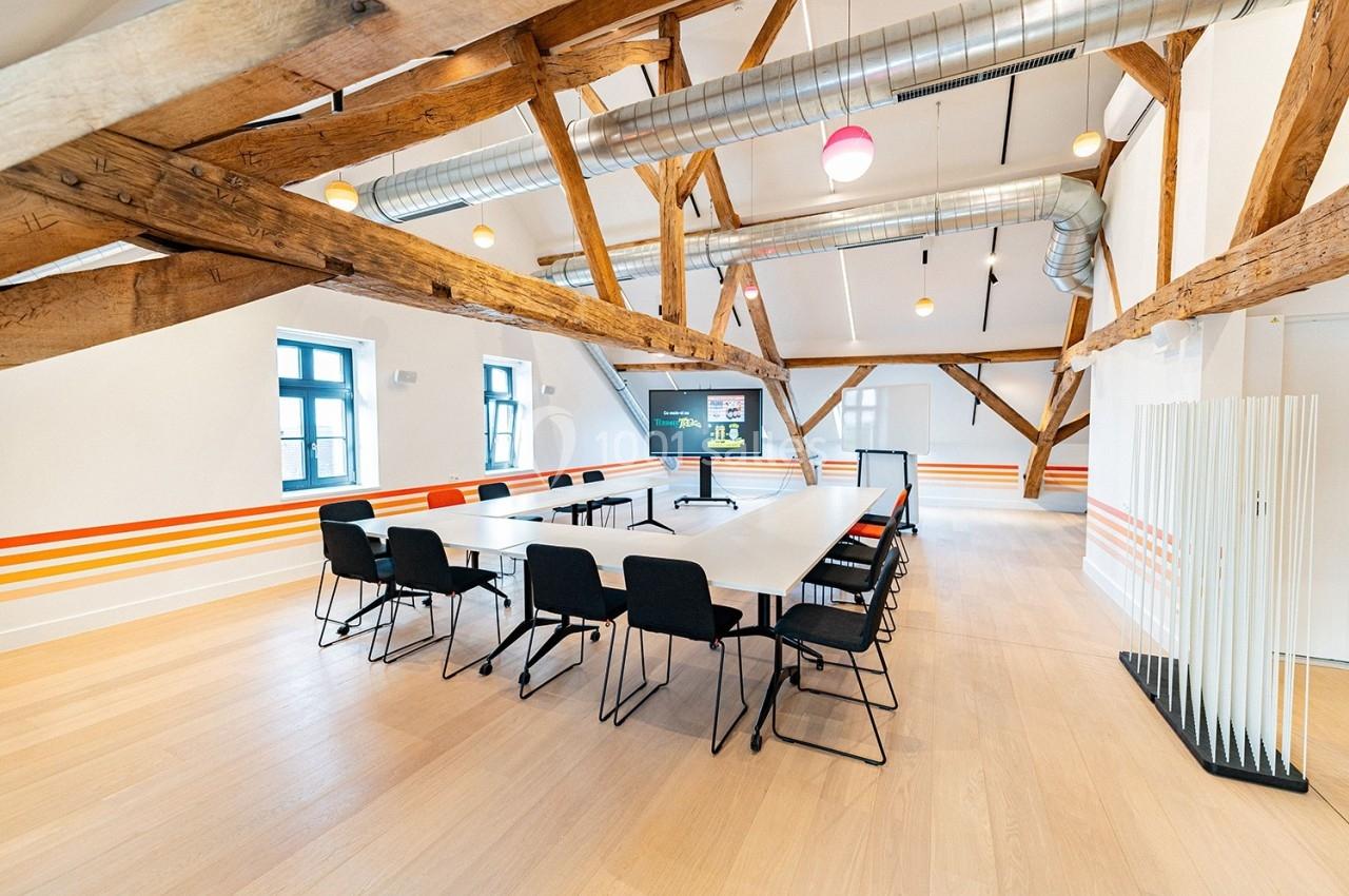 Salle de réunion lumineuse avec poutres apparentes, grande table rectangulaire, chaises noires et écran de présentation.