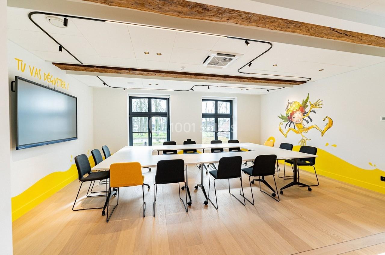 Salle de réunion lumineuse avec table en U, chaises noires et jaunes, écran mural et décorations murales colorées.